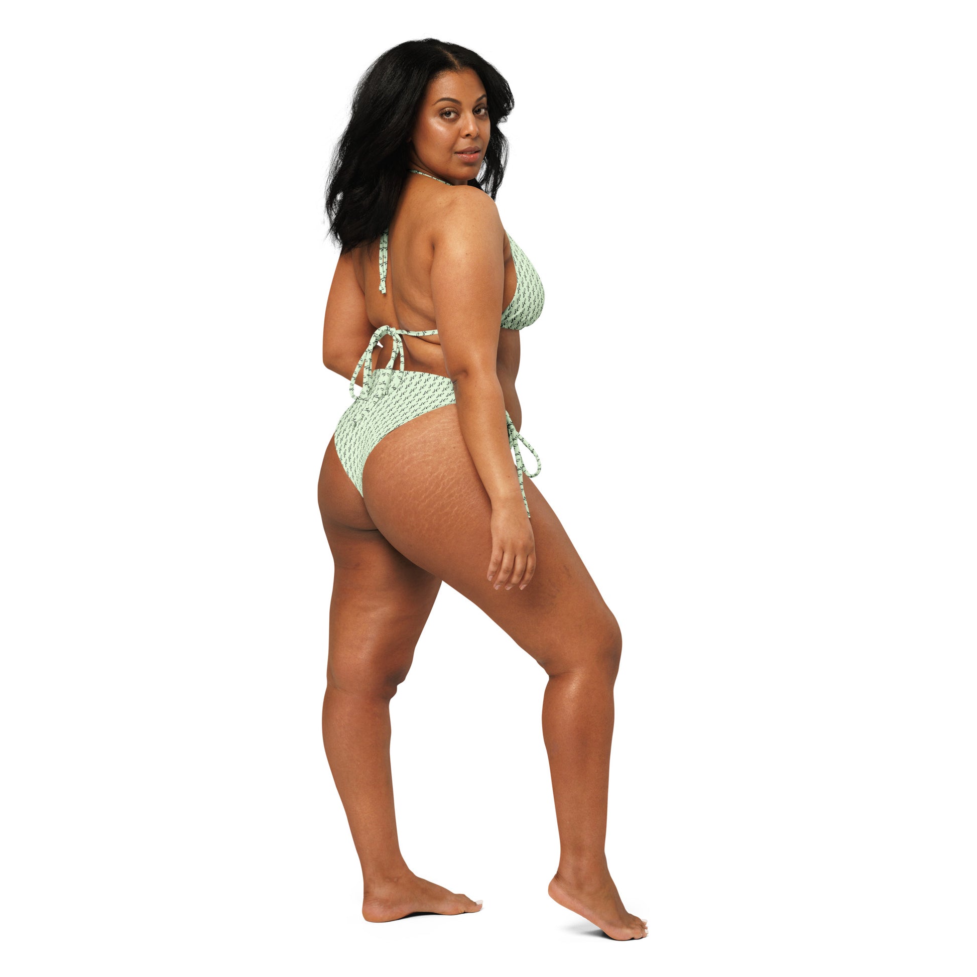 A confident woman wearing an Emerald Script Recycled String Bikini in light green with script patterns, shown from the back, highlighting the bikini's adjustable string ties.