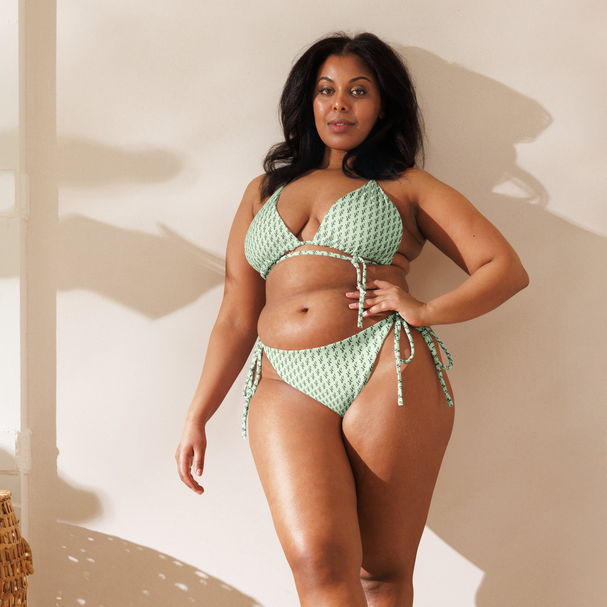 Woman wearing an Emerald Script Recycled String Bikini in a soft green patterned fabric, posing confidently indoors with natural lighting and a neutral background.