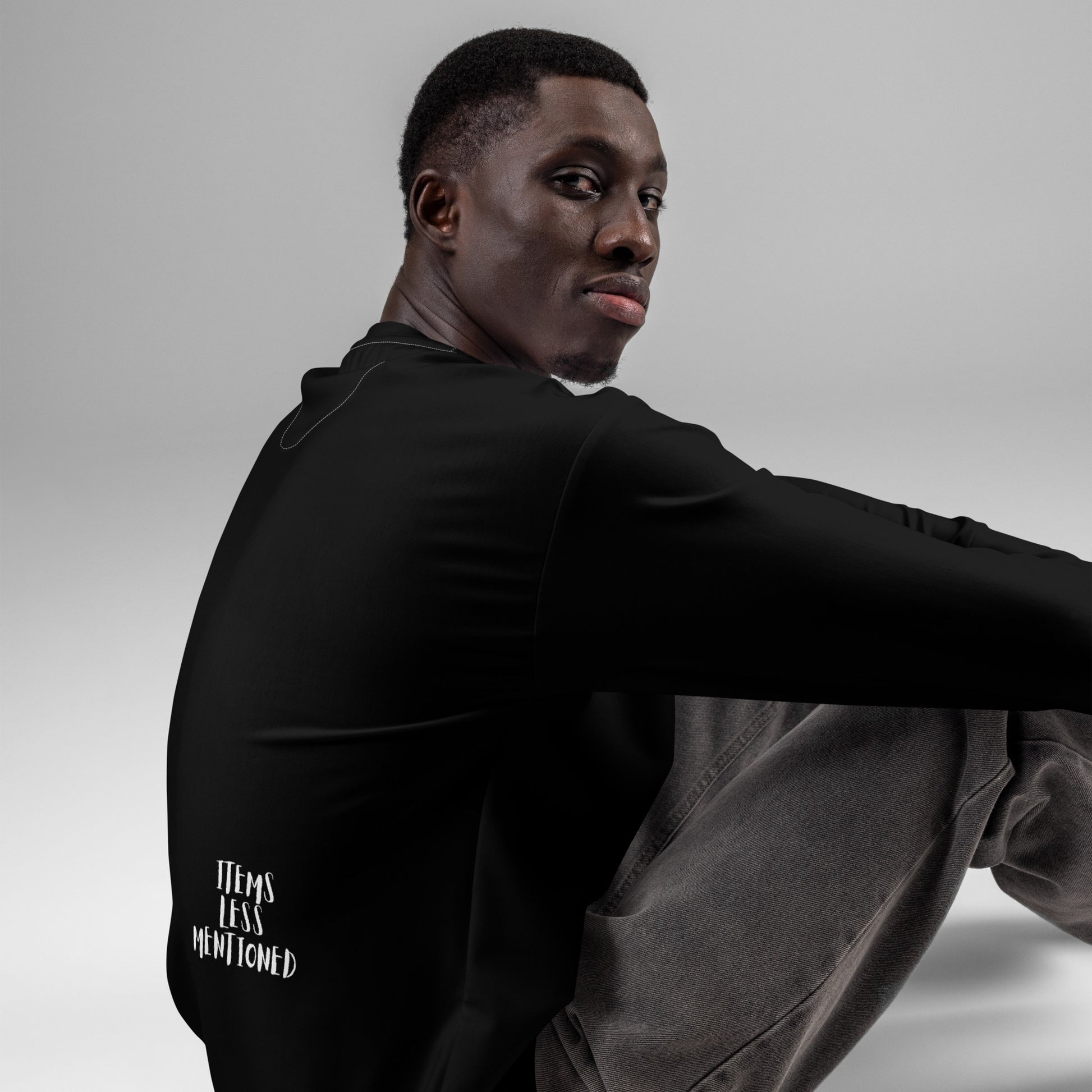 Man seated wearing a black Coffee Withdrawal Premium Sweatshirt featuring white "Items Less Mentioned" text, paired with gray pants against a plain gray background.