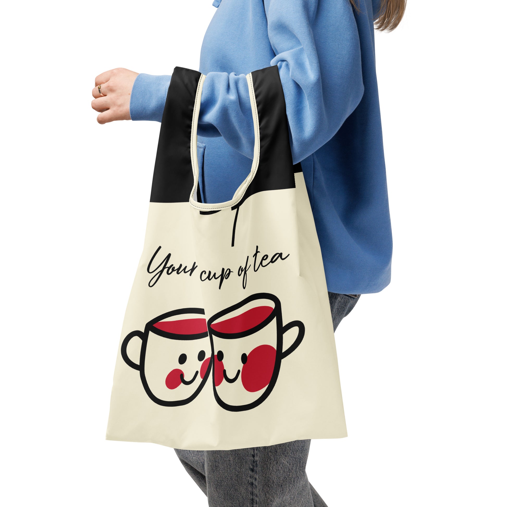 Person wearing a blue sweatshirt holding a cream and black Shopping Bag – You're My Cup of Tea with a cute illustration of two smiling tea cups, featuring faith-inspired and purpose-driven messaging.