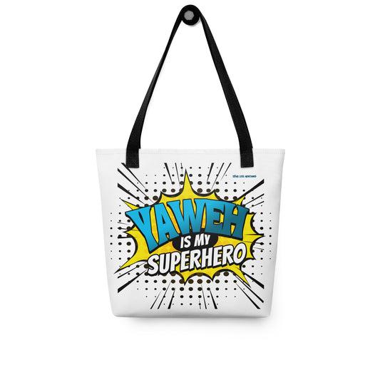 White tote bag with black handles featuring colorful comic-style text saying "Tote Bag – Yahweh Is My Superhero" in blue, yellow, and black, designed with a faith-inspired theme.