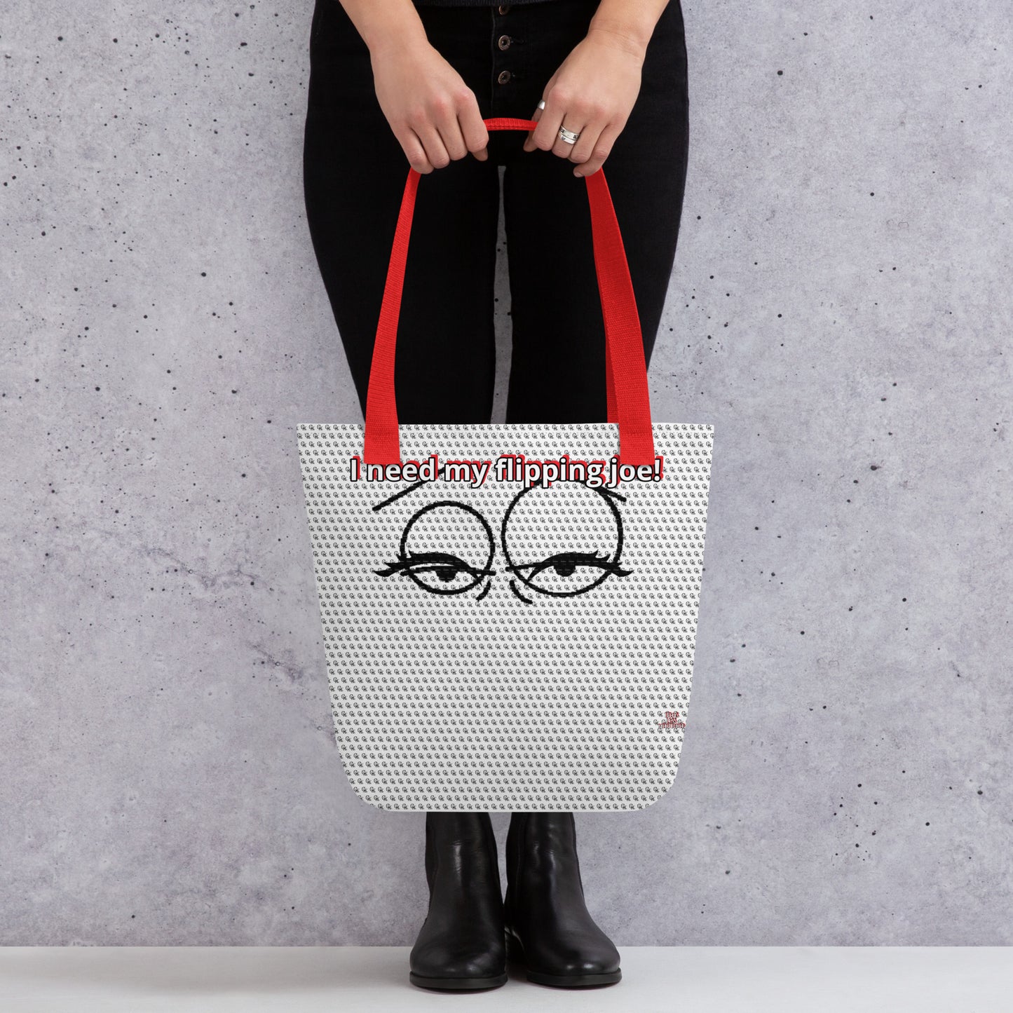 Person holding a printed tote bag with red handles featuring sleepy eyes and the phrase "I need my flipping joe!" on a light textured background, showcasing the Bag – I Need My Flipping Joe.