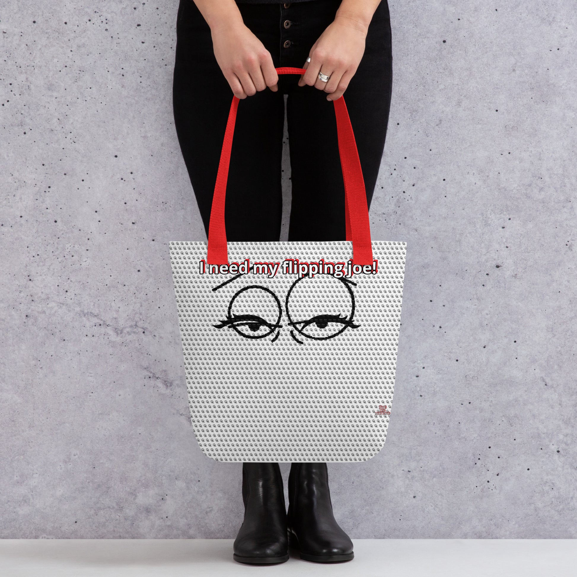 Person holding a printed tote bag with red handles featuring sleepy eyes and the phrase "I need my flipping joe!" on a light textured background, showcasing the Bag – I Need My Flipping Joe.