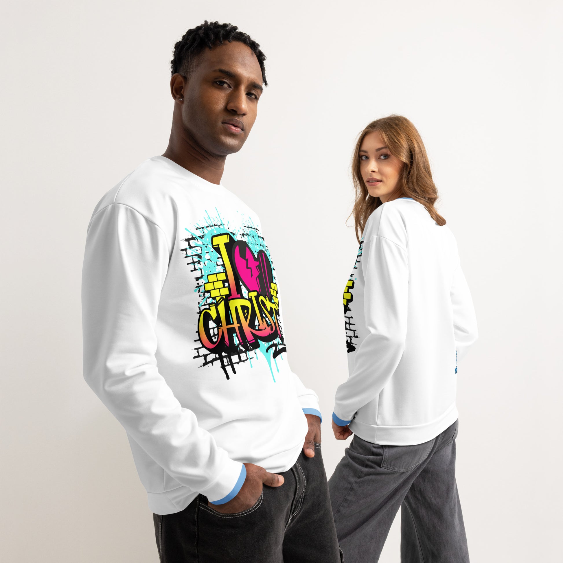 Two models wear a white Unisex Sweatshirt – I Love Christ featuring vibrant, colorful graffiti-style faith-inspired artwork, blending melanin-rich imagery with purpose-driven messaging.