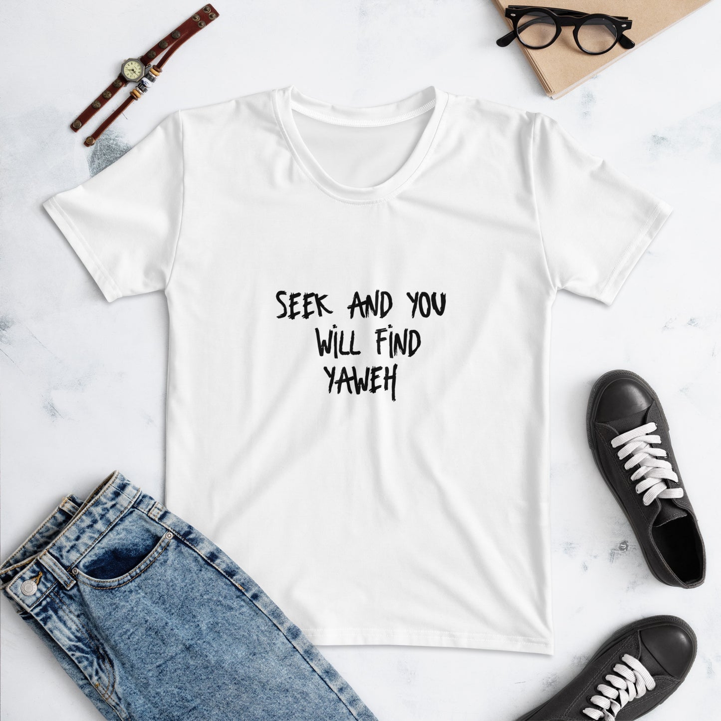White crew neck "Women's T-Shirt – Seek and You Shall Find Yahweh" with black text, styled with denim jeans, black sneakers, watch, glasses, and notebook on a light background.