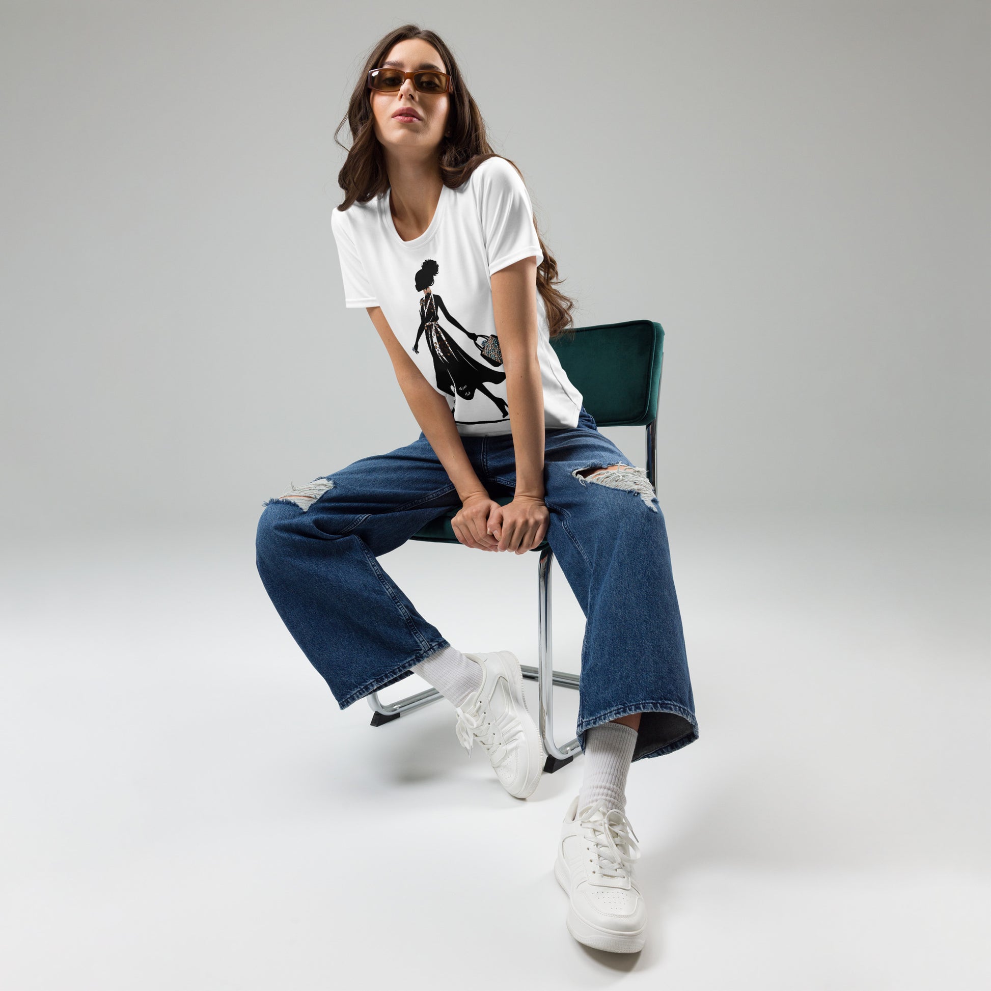 Woman wearing a white Afro Puff Strut Women's T-Shirt with black graphic design, paired with ripped blue jeans, white sneakers, and sunglasses, sitting on a chair.