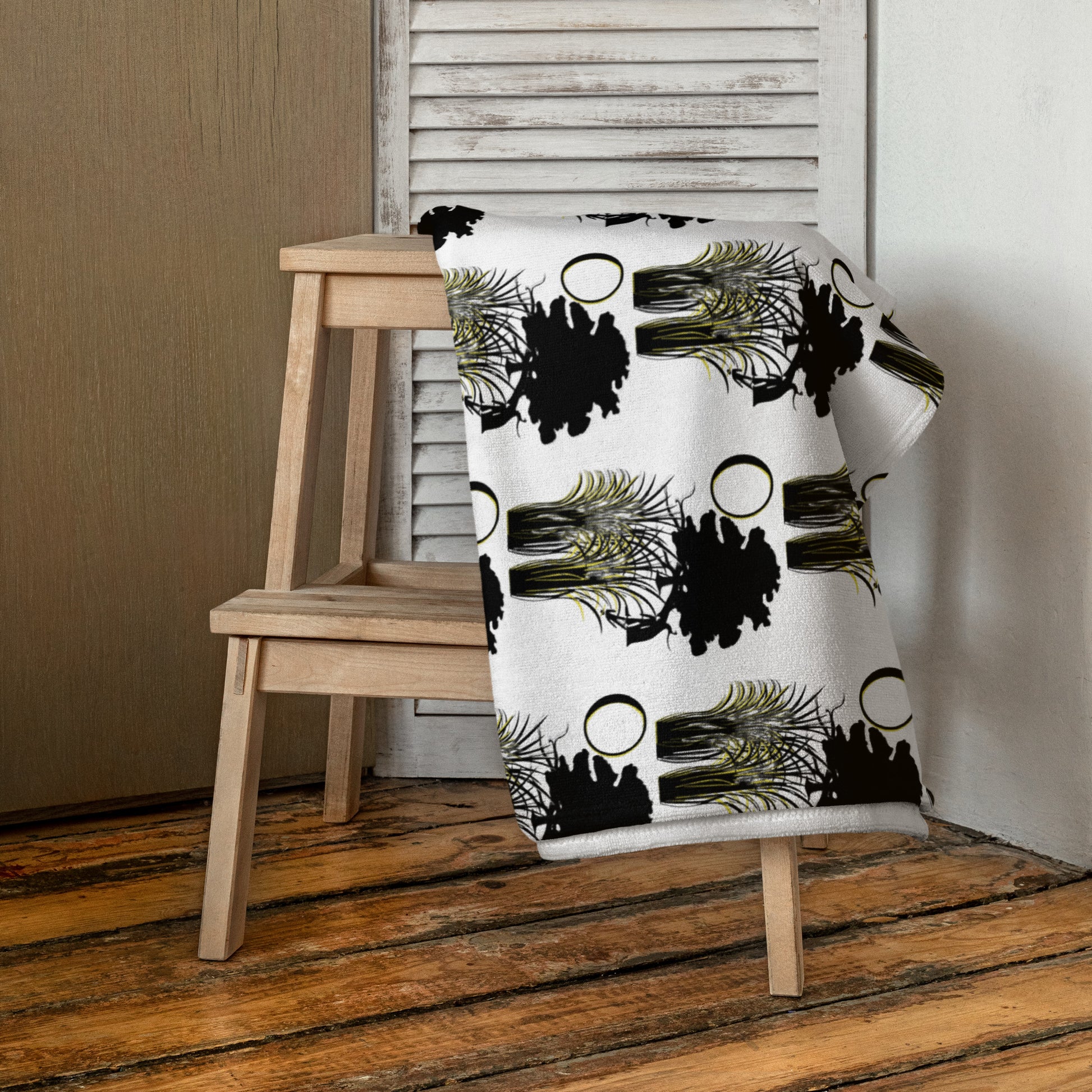 Moon & Wildgrass Towel draped over a wooden chair, featuring a white background with black silhouette trees and abstract gold accents, embodying faith-inspired and melanin-rich design.