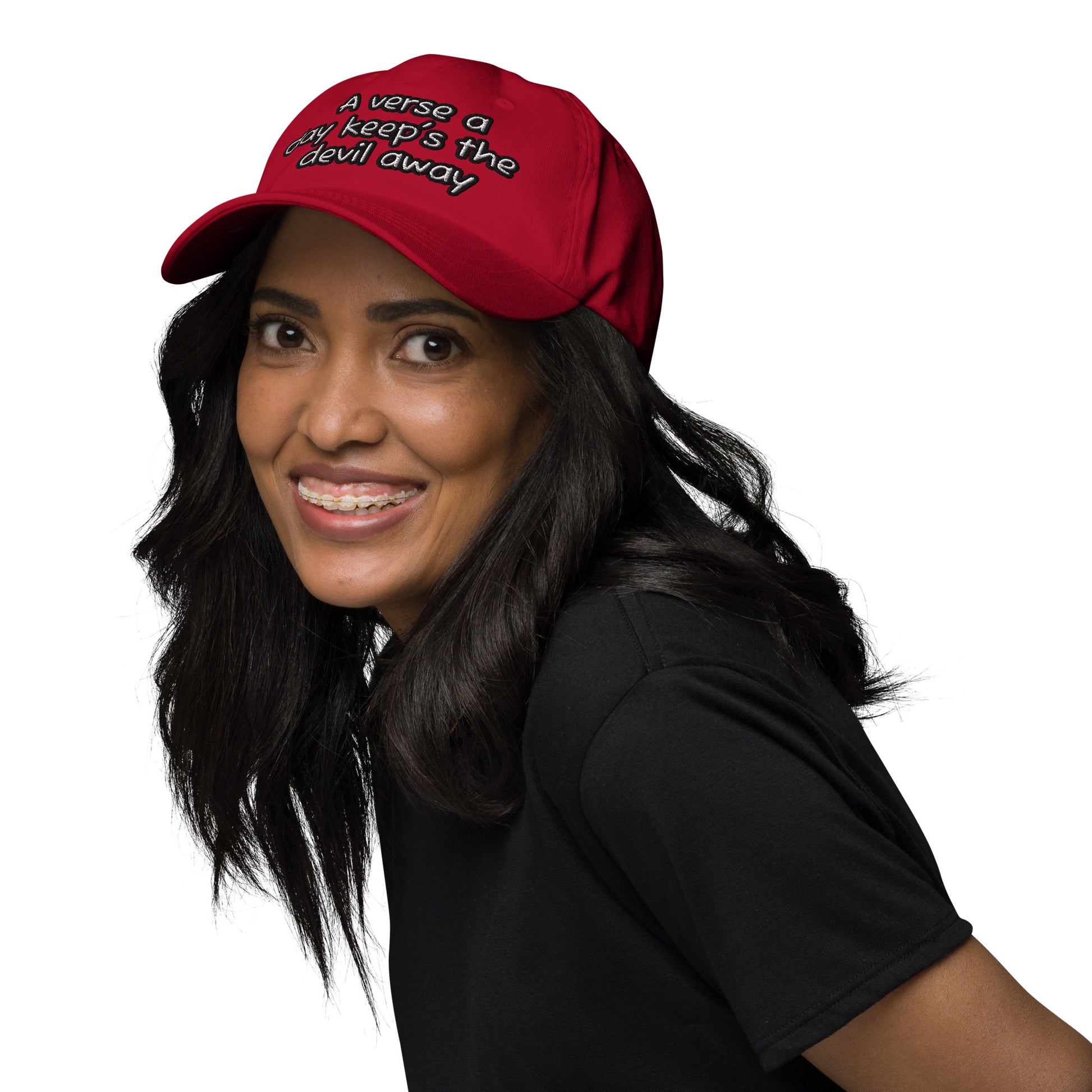 Smiling woman wearing a red Dad Hat – A Verse a Day Keeps the Devil Away with black text, styled with loose dark hair and a black shirt.