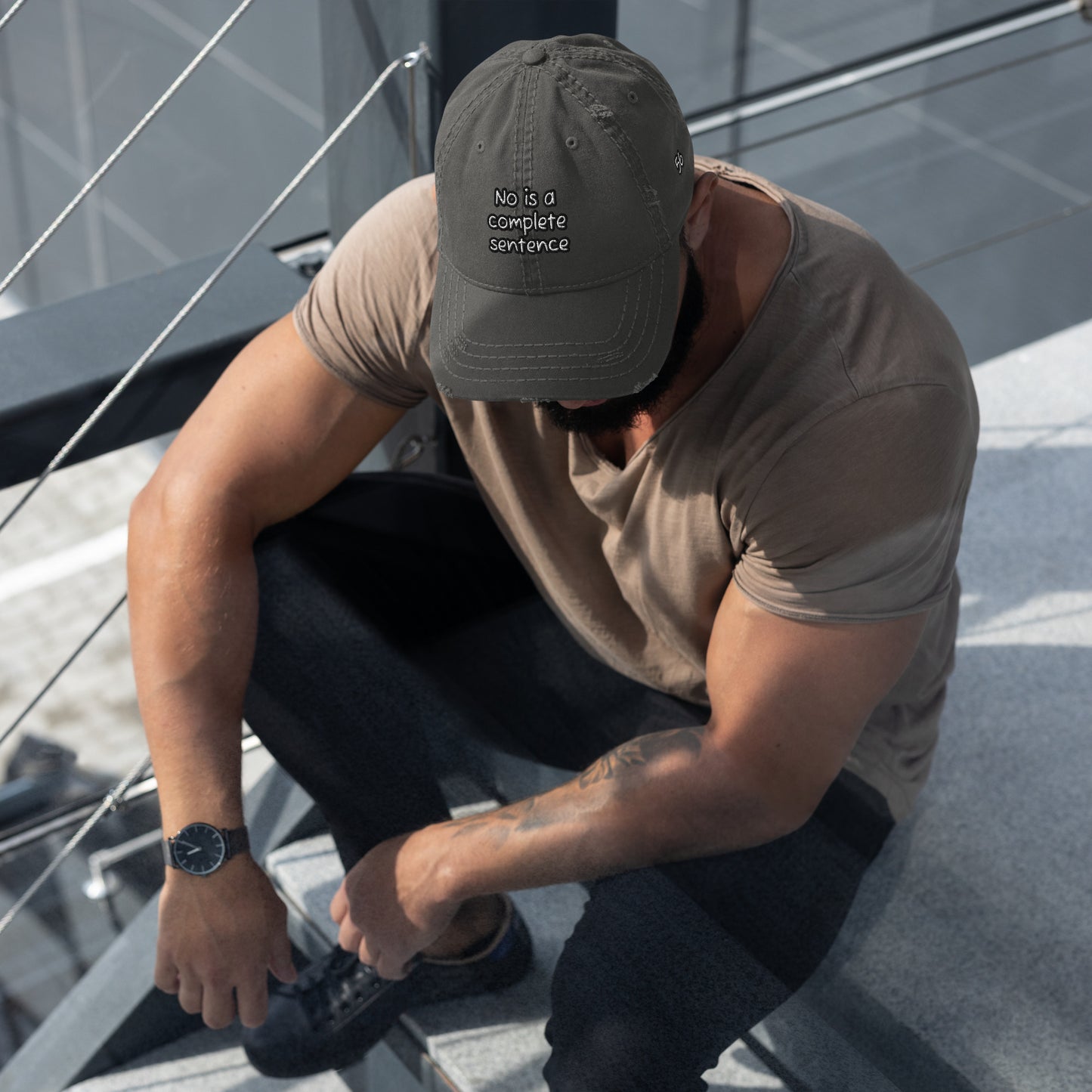 Man sitting outdoors on concrete steps wearing a charcoal grey Distressed Dad Hat – No Is a Complete Sentence with embroidered text, paired with a taupe t-shirt and black pants, casual style