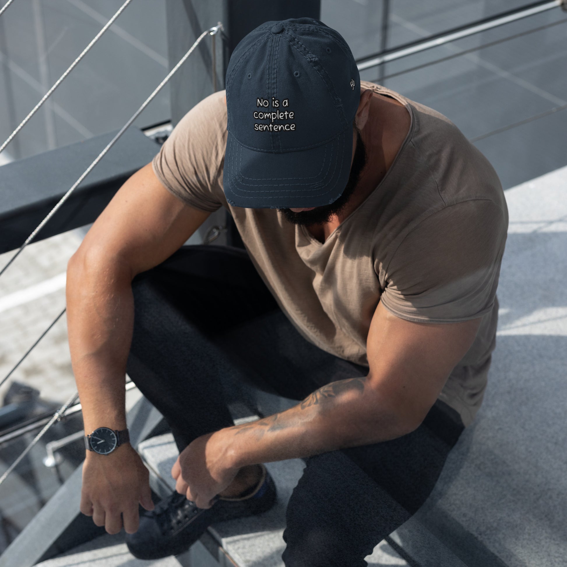 Man sitting on steps wearing a navy Distressed Dad Hat – No Is a Complete Sentence with casual brown t-shirt and black pants in outdoor urban setting