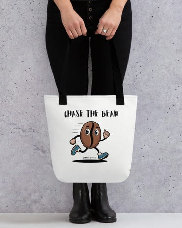 Person holding a white Tote Bag – Chase the Bean with black handles, featuring a cartoon coffee bean character running and the text "CHASE THE BEAN" above it.
