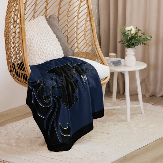 Cozy navy blue Sherpa Blanket – Moonlit Elder draped over a wicker chair with white pillow, featuring intricate designs, next to a small white table with flowers in a softly lit room.