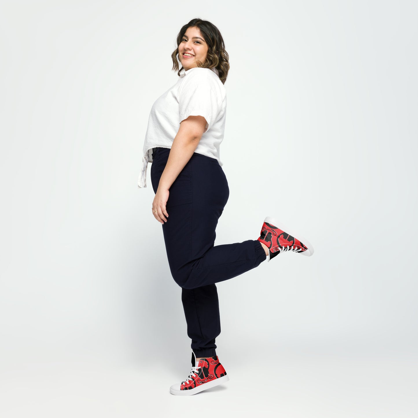 Woman smiling and posing with one leg lifted wearing colorful Women's High Top Canvas Shoes – Plug In, white short-sleeve top, and dark pants against a plain background.