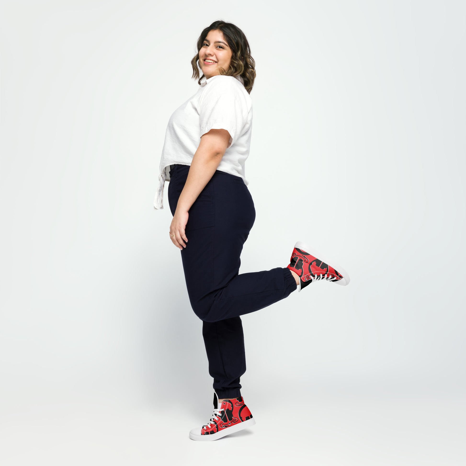Woman smiling and posing with one leg lifted wearing colorful Women's High Top Canvas Shoes – Plug In, white short-sleeve top, and dark pants against a plain background.